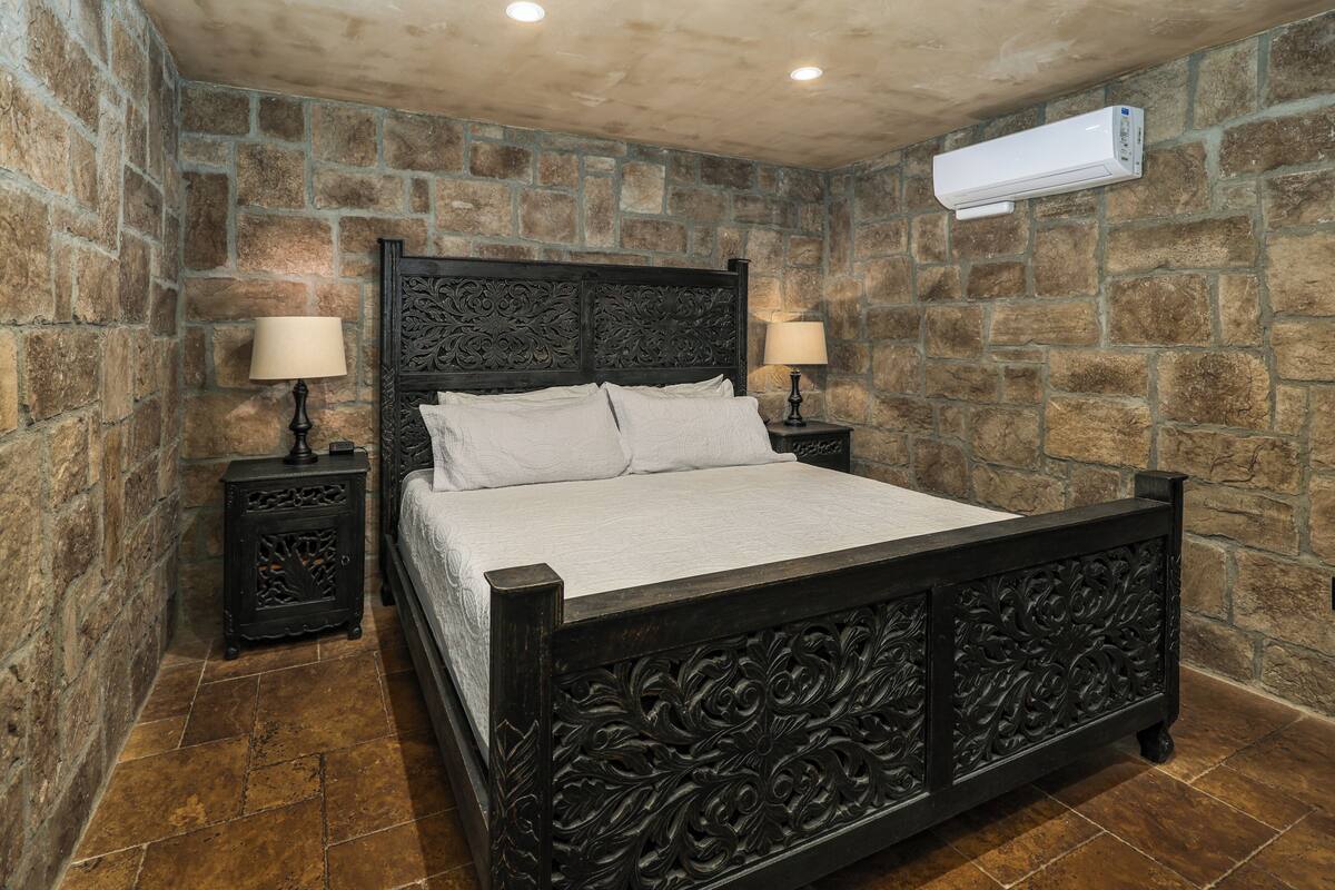 A bedroom with a stone wall, a bed with a dark headboard, two bedside tables with lamps, and an air conditioning unit on the wall.