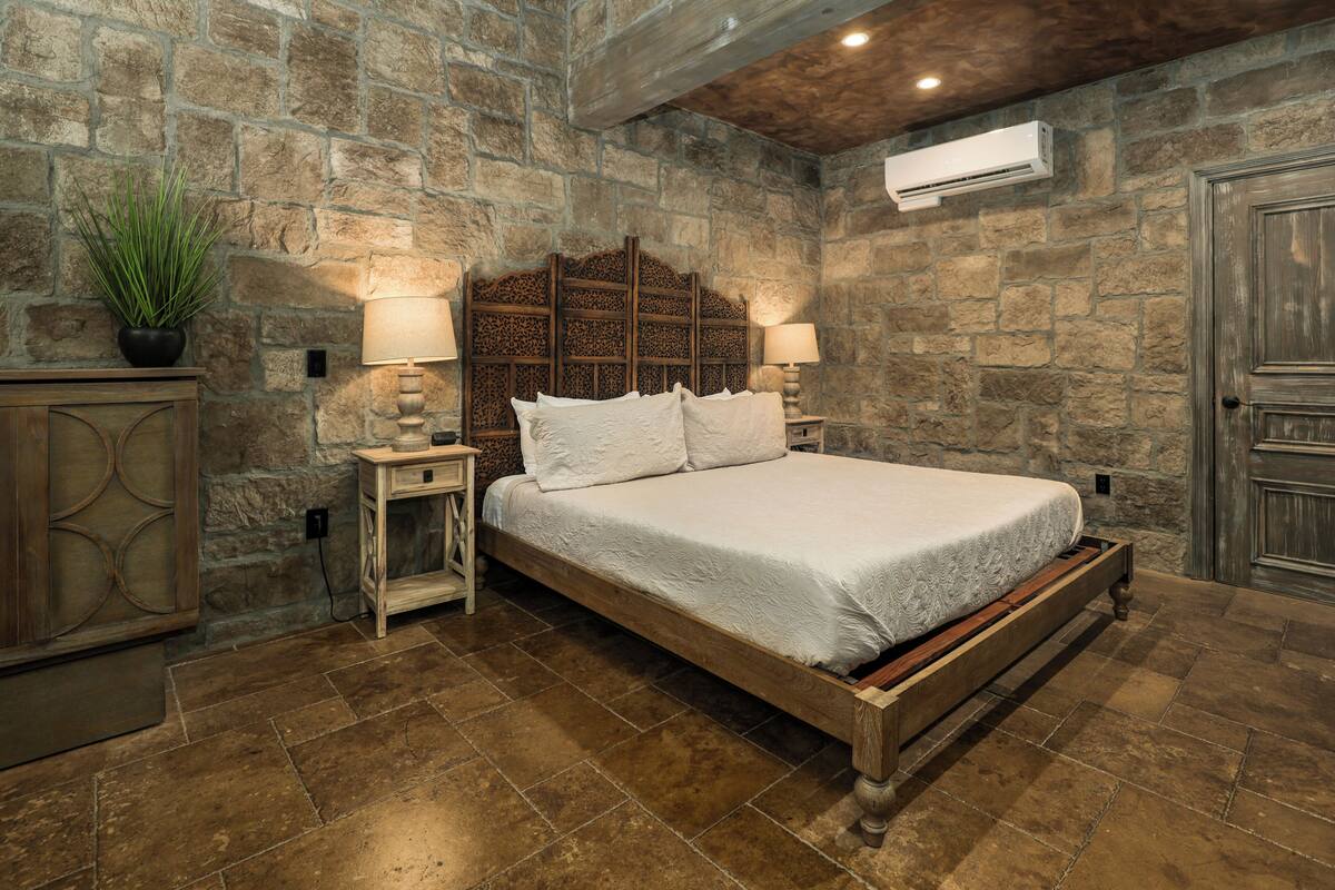 A bedroom with a stone wall, a wooden bed frame, and a nightstand with a lamp.