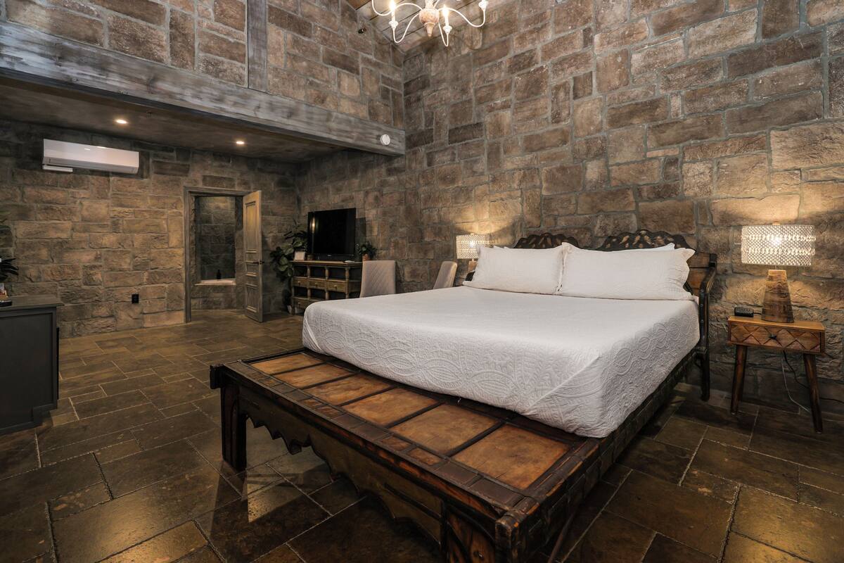 A bedroom with a stone wall, a wooden bed frame, a bench, a bedside table with a lamp, and a television.