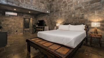 Un dormitorio con pared de piedra, estructura de cama de madera, un banco, mesita de noche con lámpara y televisor.