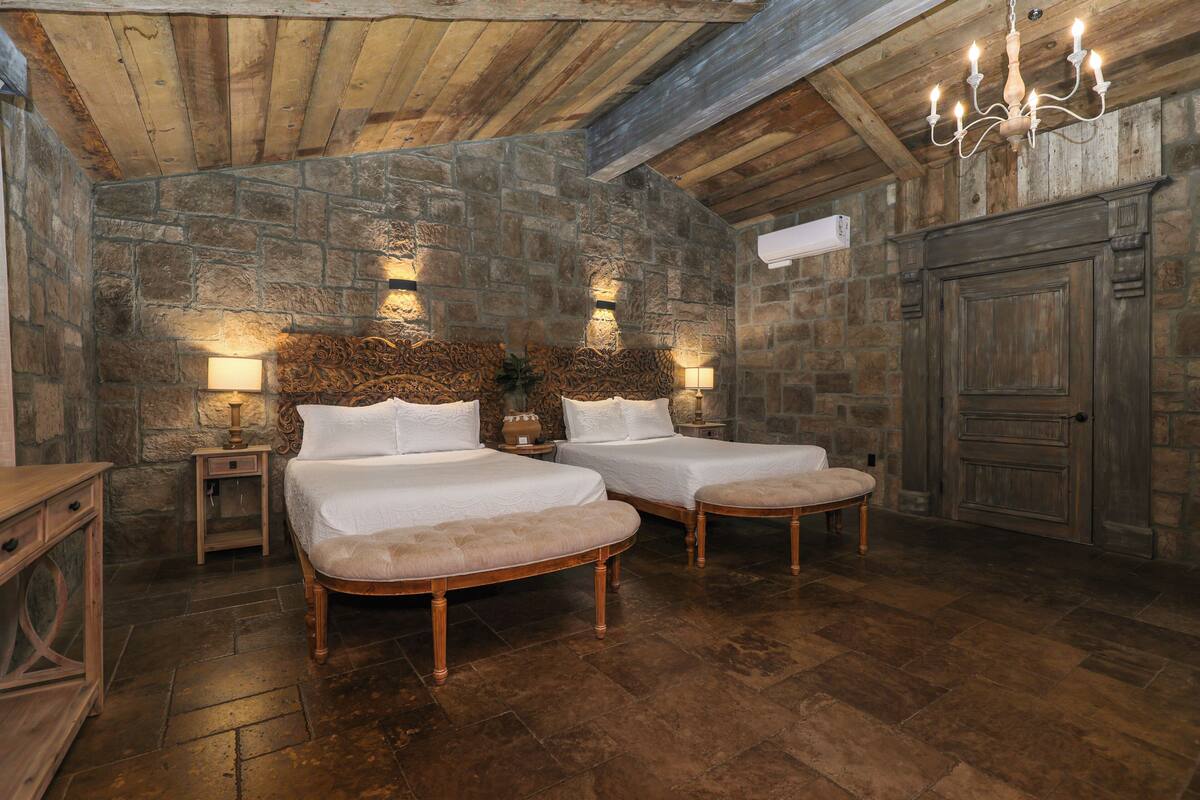 A rustic bedroom with two beds, stone walls, wooden beams, and a chandelier.