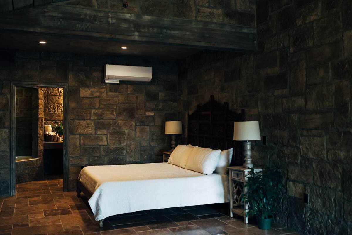 A bedroom with a stone wall, a bed with white linens, two bedside tables with lamps, and an air conditioning unit.
