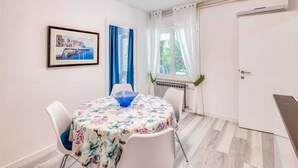 A dining area with a round table covered in a floral tablecloth, two white chairs, a framed picture on the wall, and a window with curtains. - Apartment Moracea near the beach (Njivice)