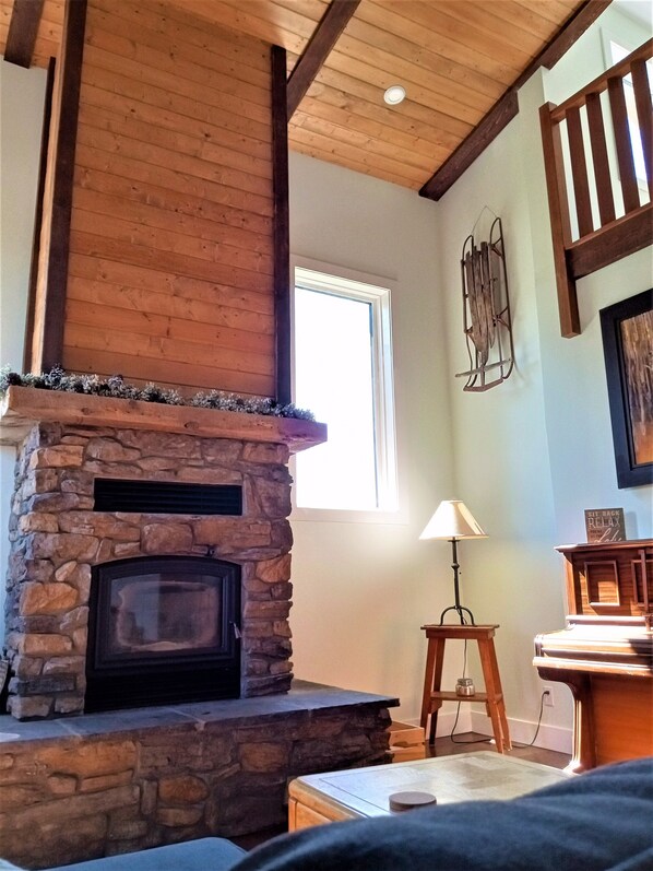 A stone fireplace with a wooden mantel, a window with blinds, a lamp, and a rustic wooden ceiling. - Perkin Lodge (Invermere)