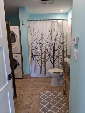 A compact bathroom with a washing machine, toilet, and shower curtain with a tree design. - Perkin Lodge (Invermere)