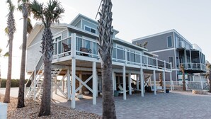 A two-story beachfront house with a covered deck, palm trees, and a pebble pathway.