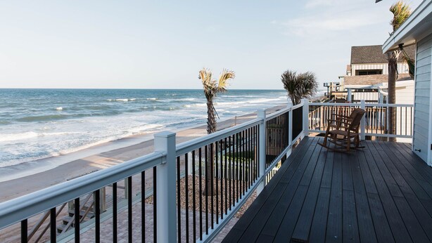 A wooden deck with a rocking chair, overlooking a beach with waves and a clear sky.