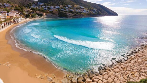A coastal view with a beach, clear turquoise waters, and a hillside with buildings.