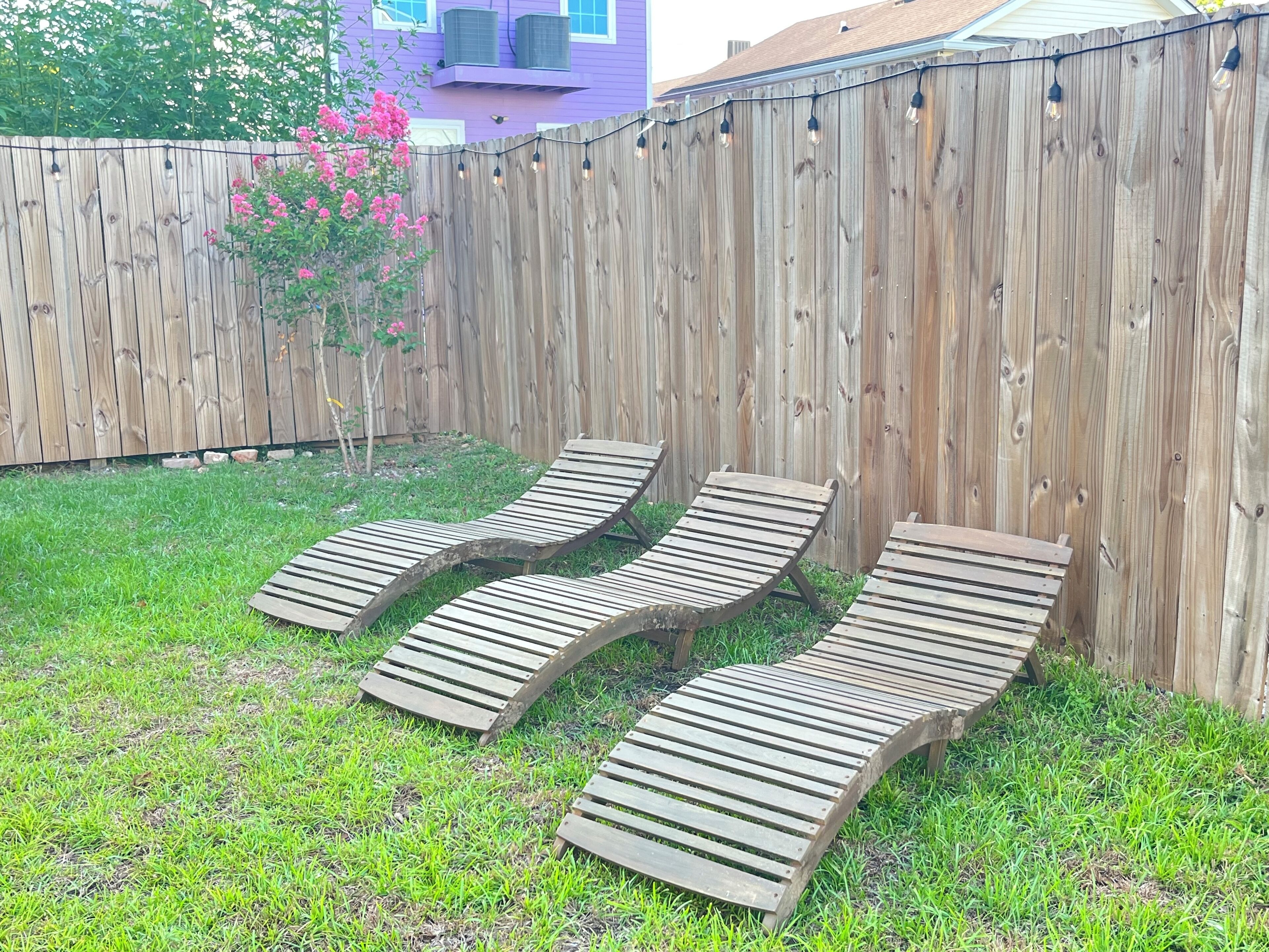 A backyard with a wooden fence, three wooden lounge chairs, and a small tree with pink flowers.