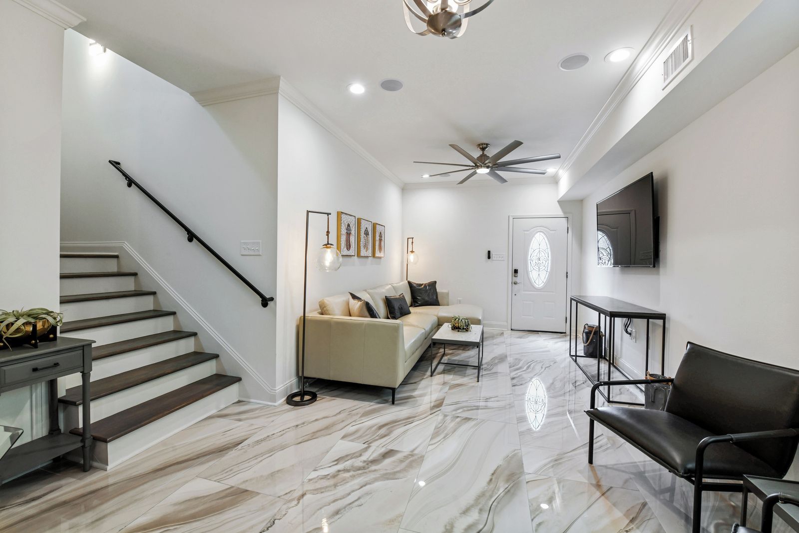 A modern living room with a marble floor, a sofa, a coffee table, a black leather chair, a TV mounted on the wall, and a staircase with a black railing.