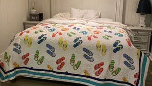 Ein Schlafzimmer mit einem Bett, das mit einer farbenfrohen Stegdecke bezogen ist, einem Deckenventilator, einem Nachttisch mit Lampe und einer Kommode.