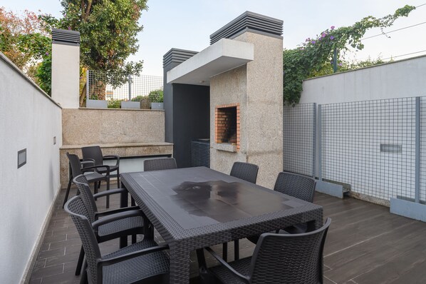 An outdoor patio with a rectangular table and six chairs, a small brick oven, and a stone wall.