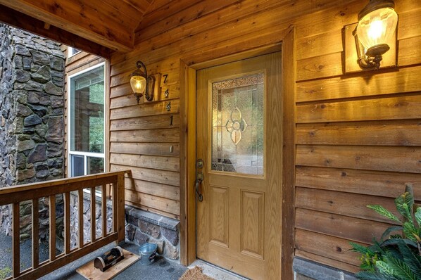 Entrada a una cabaña de madera con un muro de piedra, una puerta de madera con un panel de vidrio y dos luces fijadas en la pared.