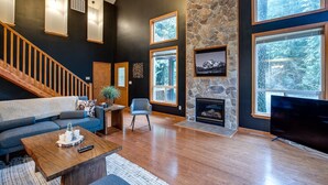 Amplia sala de estar con chimenea de piedra, ventanas grandes, escalera de madera y televisor de pantalla plana.