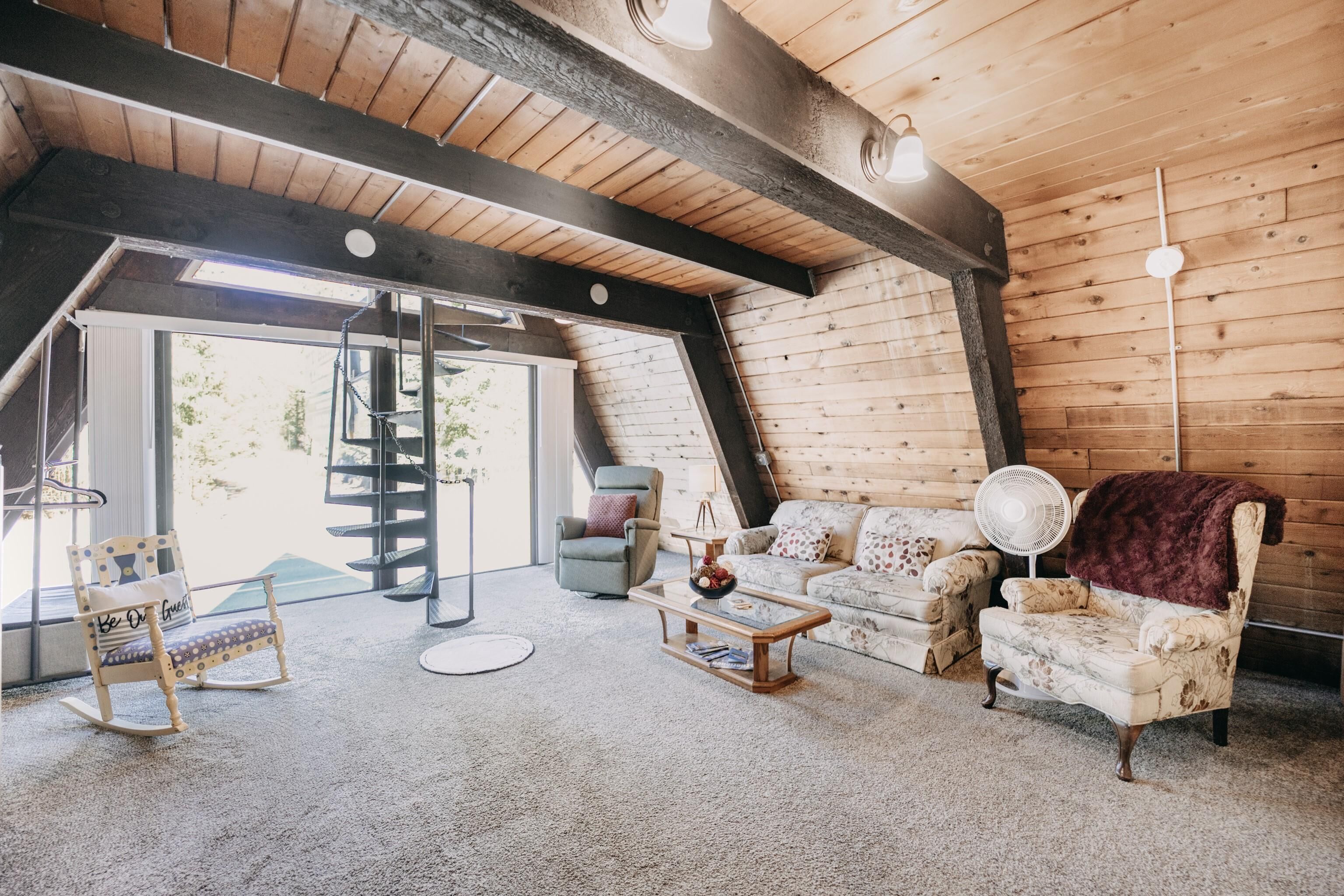 A cozy loft-style room with a wooden ceiling, a large window, a sofa, a rocking chair, and a fan.