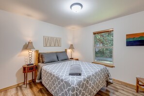 A bedroom with a bed, two bedside tables, a window with a view of trees, and a painting on the wall. - 61MBR - Hot Tub - Wi-Fi - Pets Ok - Sleeps 6 (Deming)