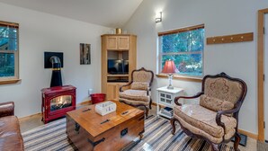 A cozy living room with a red wood-burning stove, a television, a wooden cabinet, two patterned chairs, a wooden coffee table, and a window with a view of trees. - 61MBR - Hot Tub - Wi-Fi - Pets Ok - Sleeps 6 (Deming)