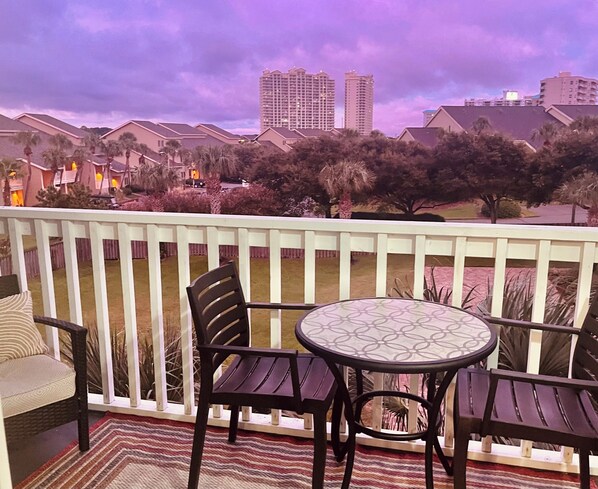 A balcony with a round table and chairs, overlooking a residential area with palm trees and buildings. - Clean & Ready for Guests! Recently Remodeled, Steps to the Beach! (Miramar Beach)