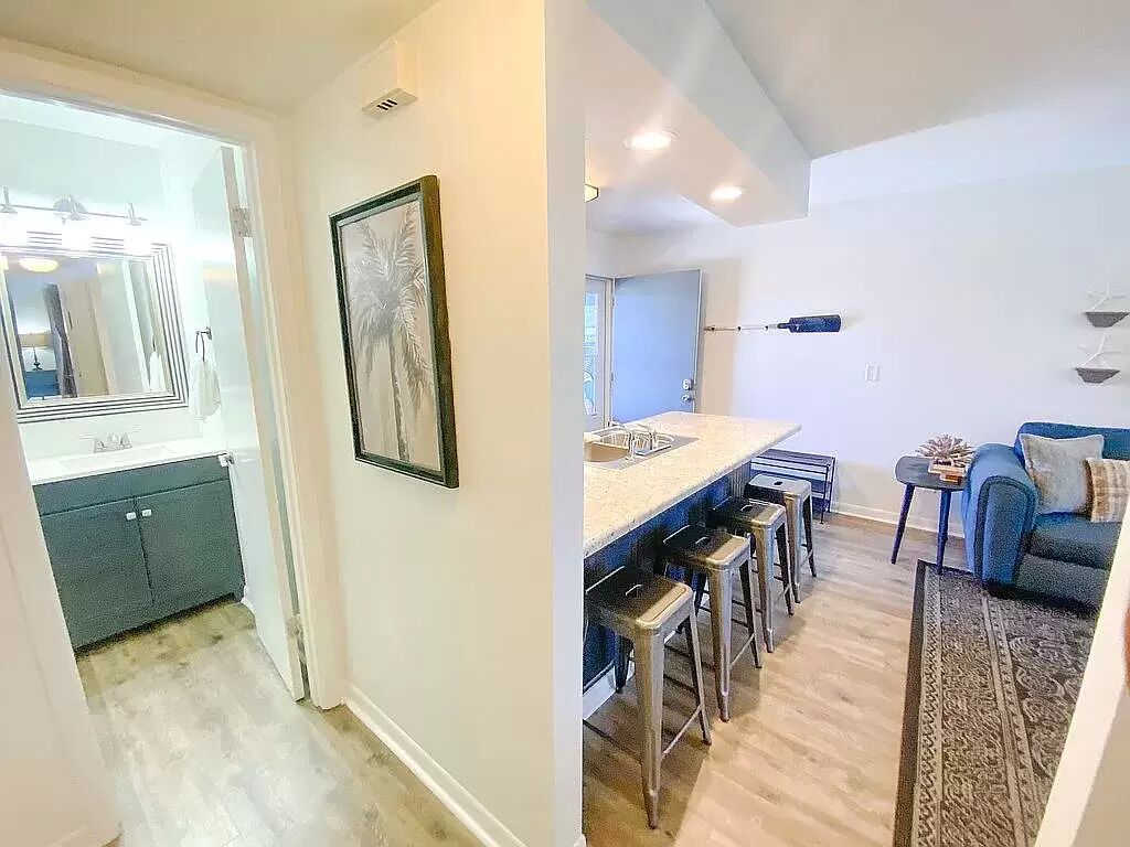 Una cocina moderna con una isla central y taburetes, una zona de comedor con un sofá azul y una alfombra estampada, y un pasillo con un espejo y obras de arte enmarcadas.