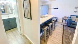 A modern kitchen with a central island and bar stools, a dining area with a blue sofa and a patterned rug, and a hallway with a mirror and framed artwork. - Clean & Ready for Guests! Recently Remodeled, Steps to the Beach! (Miramar Beach)