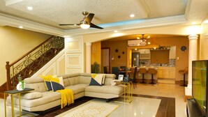 Uma sala de estar moderna com sofá seccional, uma área de jantar com banquetas, uma cozinha com balcão e uma escadaria com corrimãos decorativos.
