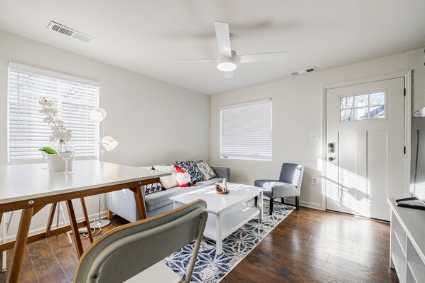 A modern living room with a white sofa, a coffee table, and a dining table. There is a ceiling fan, a door with a window, and a small plant on the windowsill.