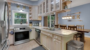A modern kitchen with a built-in oven, dishwasher, and a large island with a sink. There is a dining area with wooden chairs and a table. - Lake Hartwell Retreat, Clemson Football Weekends, 4BR + Private Dock (Westminster)