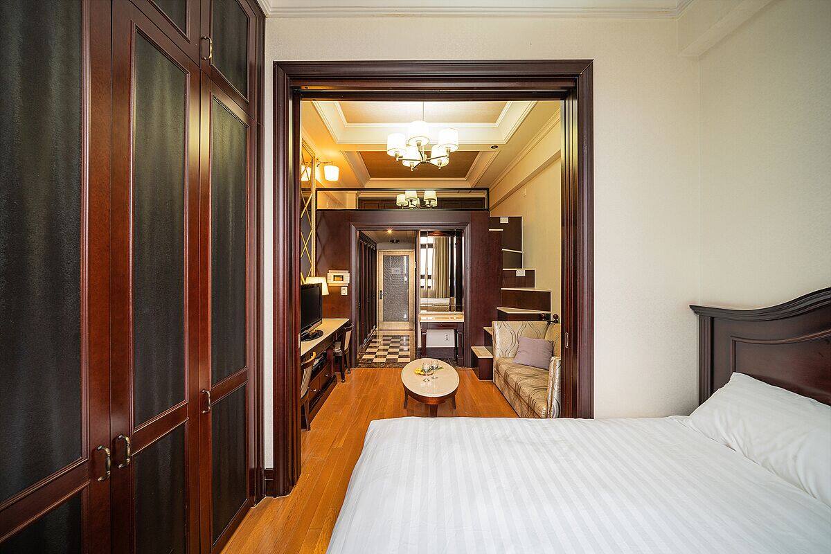 Ein Hotelzimmer mit einem großen Bett, einem kleinen runden Tisch und einer Couch. Ein Flur führt zu einem Badezimmer und einer Treppe.