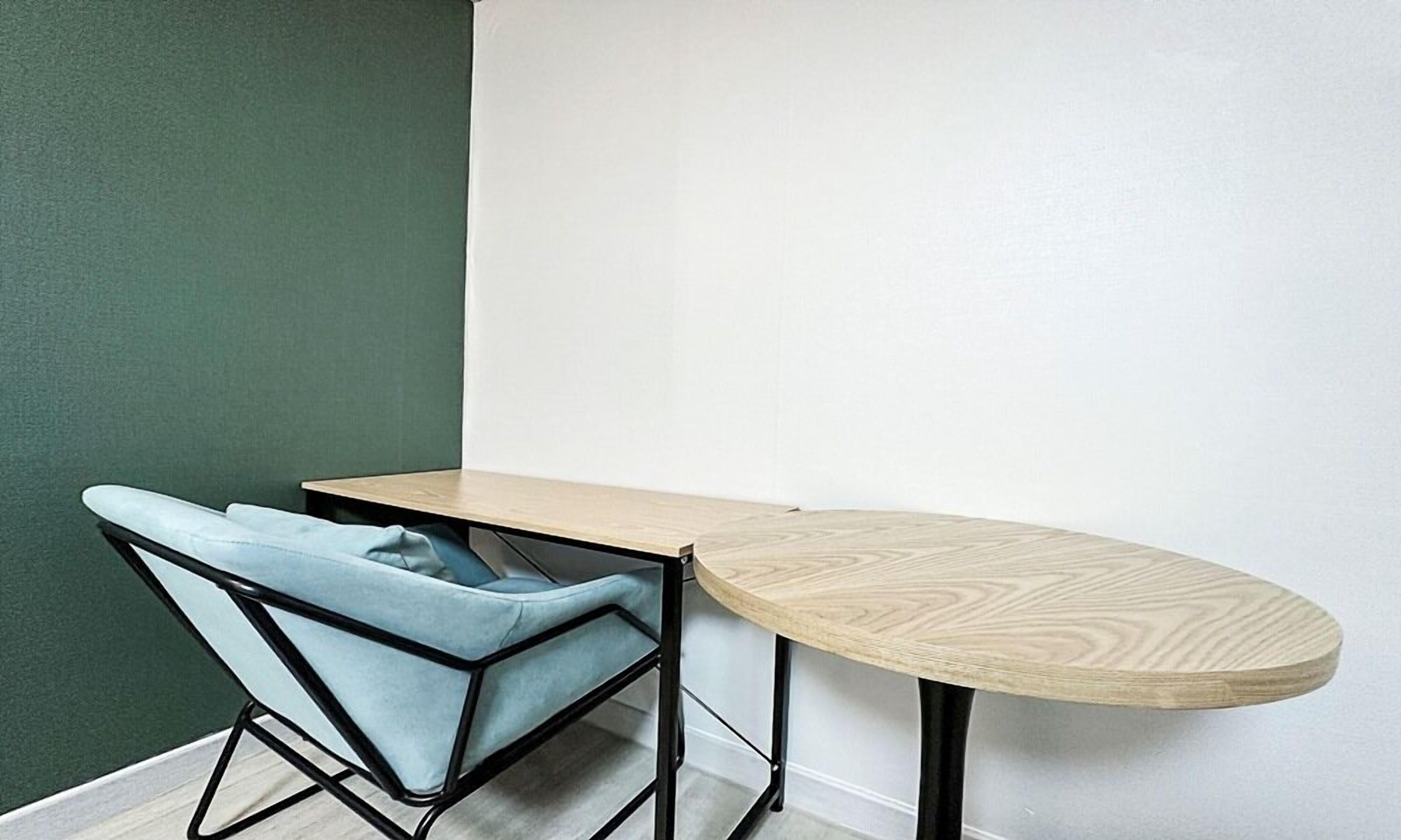 Une pièce d’angle avec un mur vert, un mur blanc, une table ronde en bois et une chaise bleue à la structure géométrique.