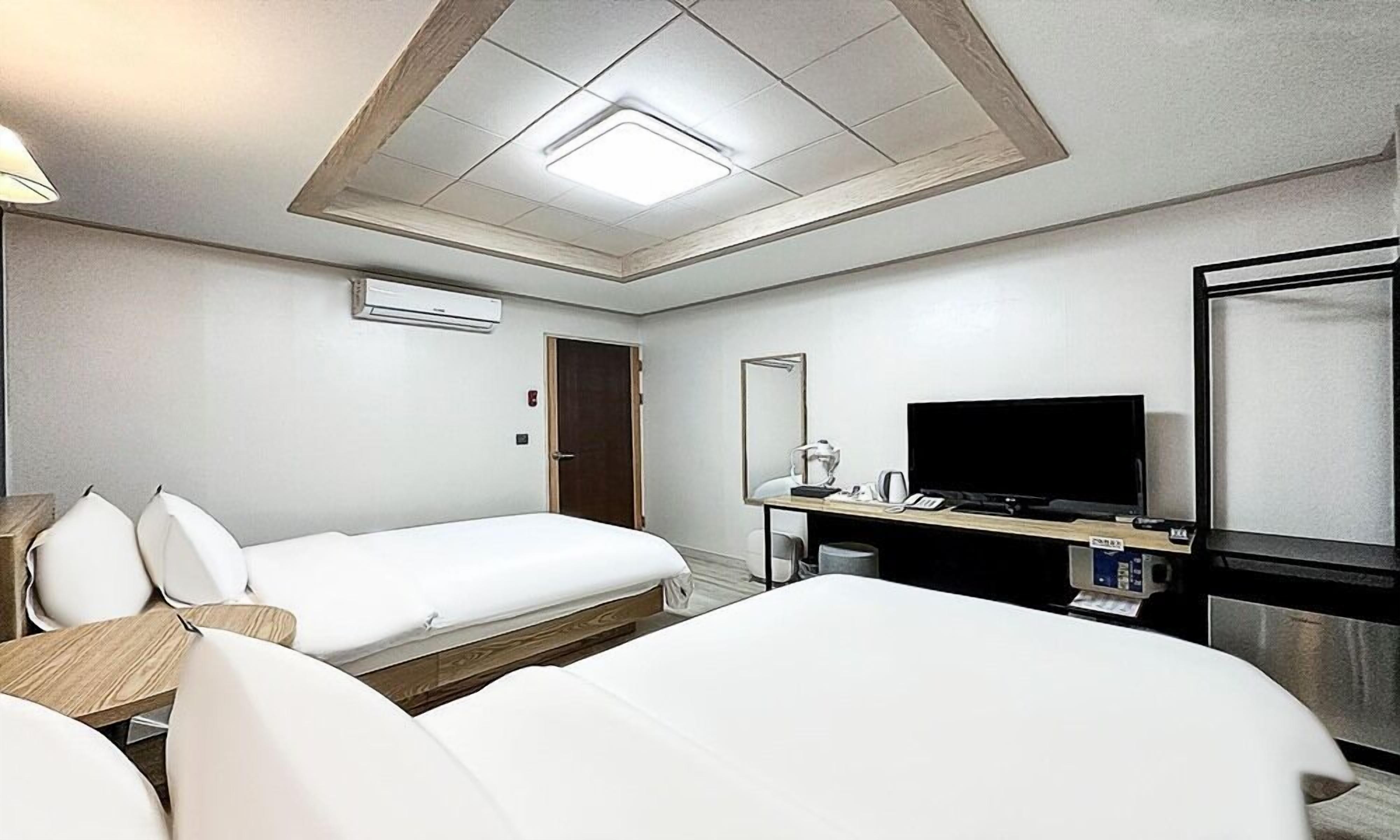 A hotel room with two beds, a desk with a flat-screen TV, a mirror, and an air conditioning unit.