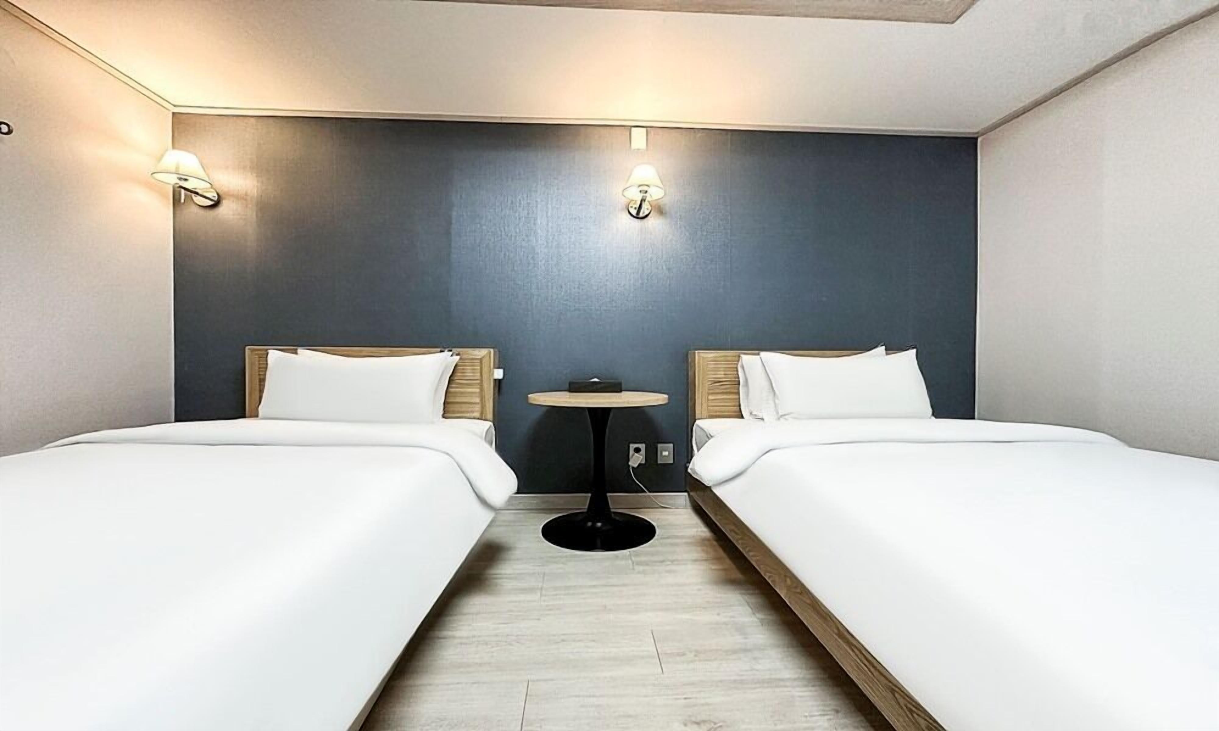 Habitación de hotel con dos camas, una mesita redonda y dos lámparas fijadas a la pared.