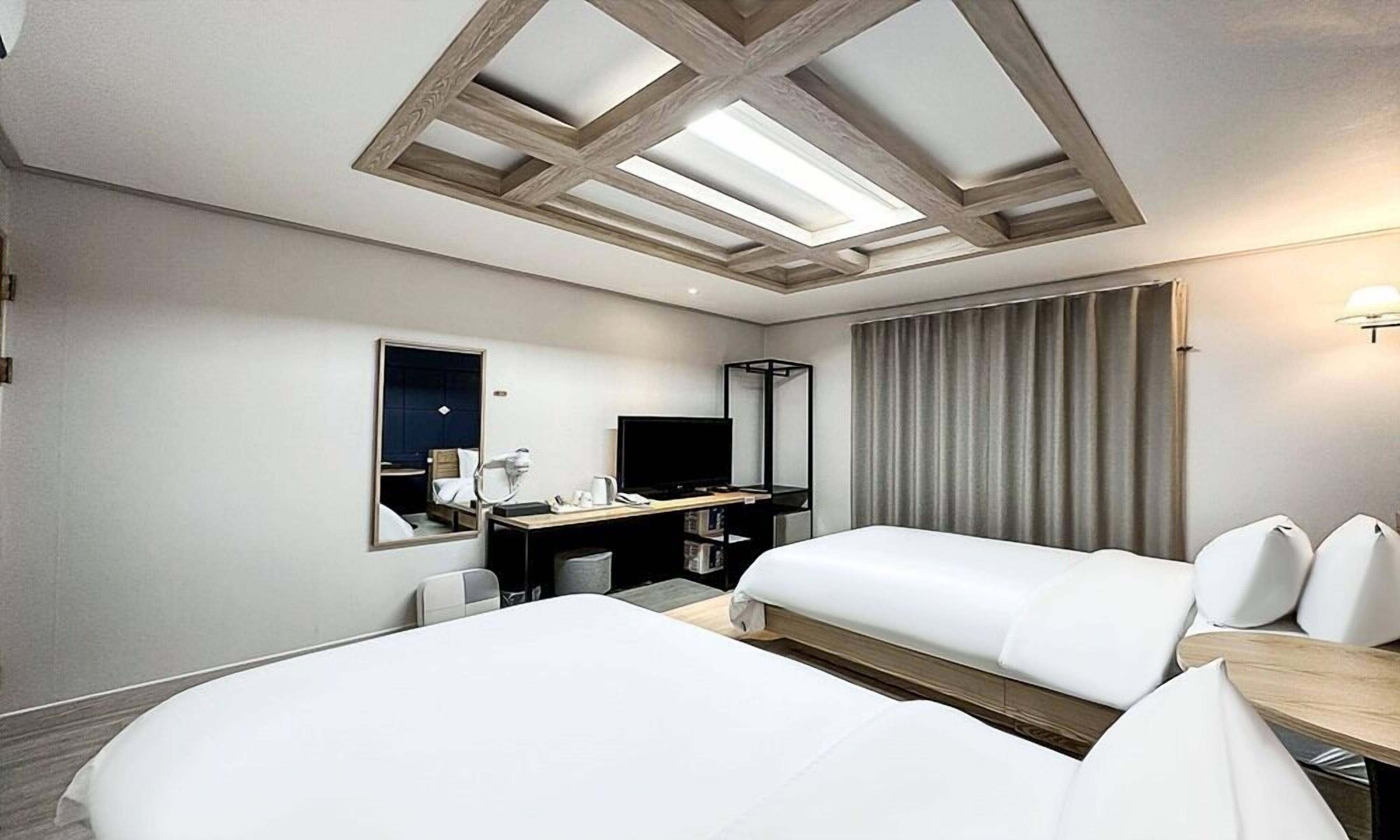 A hotel room with two beds, a desk with a computer, a TV, and a large window with curtains.