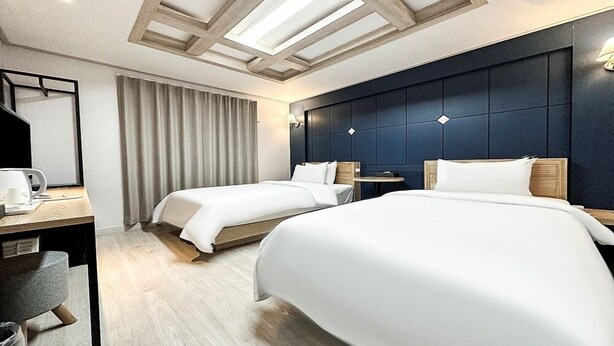A hotel room with two beds, a desk with a chair, a small table, and a large window with curtains. - Gumi Time Hotel (Gumi)