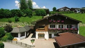 Un edificio de estilo chalet con balcón, rodeado de exuberante vegetación y un muro de piedra.