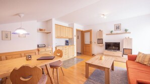 Ein modernes Wohnzimmer mit Holzesstisch, Stühlen, einer Couch und einem Fernseher. Es gibt eine Küchenzeile mit Holzschränken und einer Kaffeemaschine.