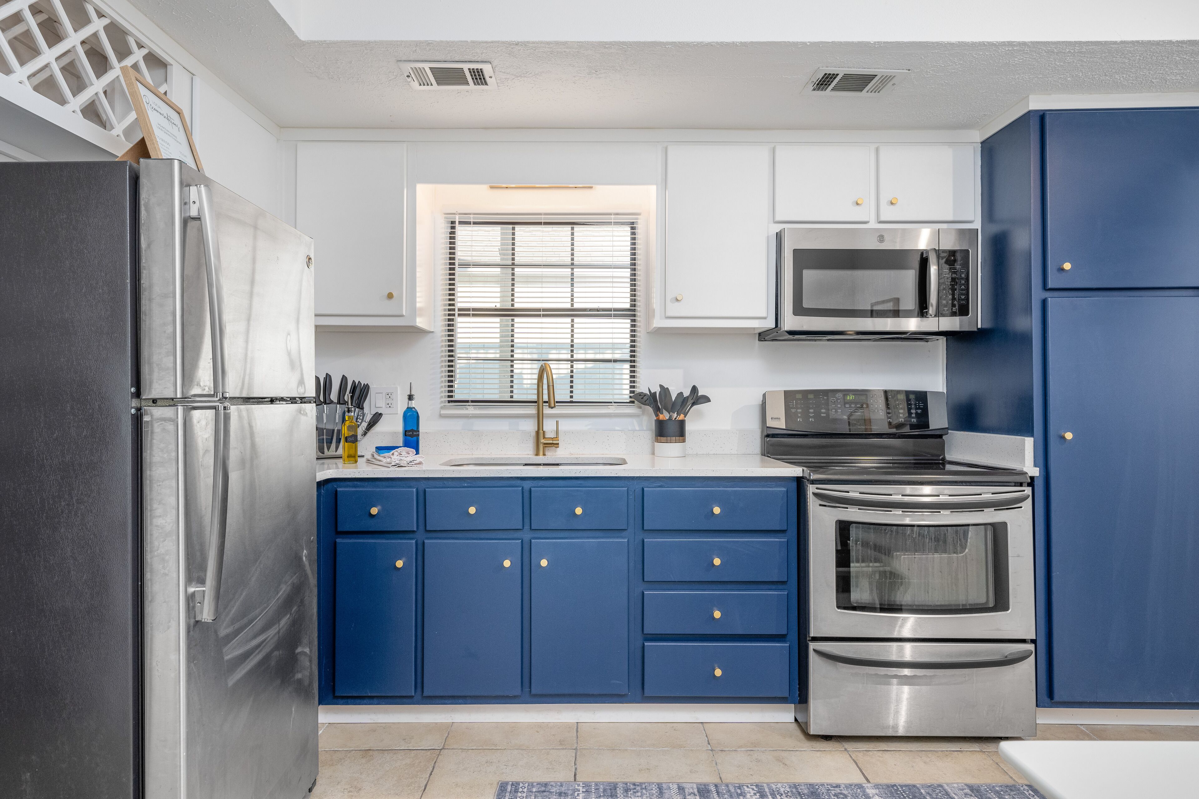 Una cocina moderna con electrodomésticos de acero inoxidable, armarios azules y un ventanal con persianas.