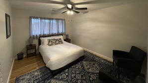 Um quarto com cama, ventilador de teto, mesa de cabeceira, cadeira e tapete estampado.