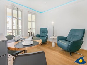 A modern room with a wooden dining table, two teal chairs, a white floor lamp, and a view of the outside through large windows.