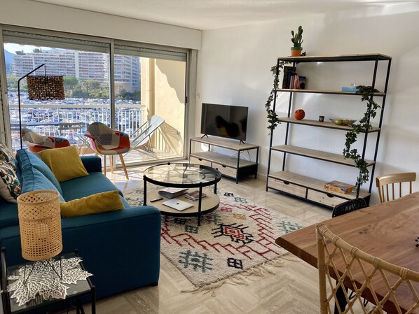 A modern living room with a blue sofa, a glass coffee table, a TV, and a bookshelf.