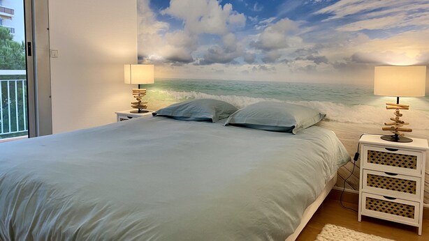 Une chambre à coucher avec un lit, deux tables de chevet avec des lampes, une fresque murale représentant une scène de plage et une fenêtre donnant sur des immeubles.