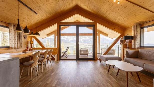 A spacious loft-style room with a wooden ceiling, a dining area with chairs and a table, a sofa, and a large sliding glass door leading to a balcony with a mountain view. - Chalet with panoramic sauna (Schladming - Pichl, Ennstal)