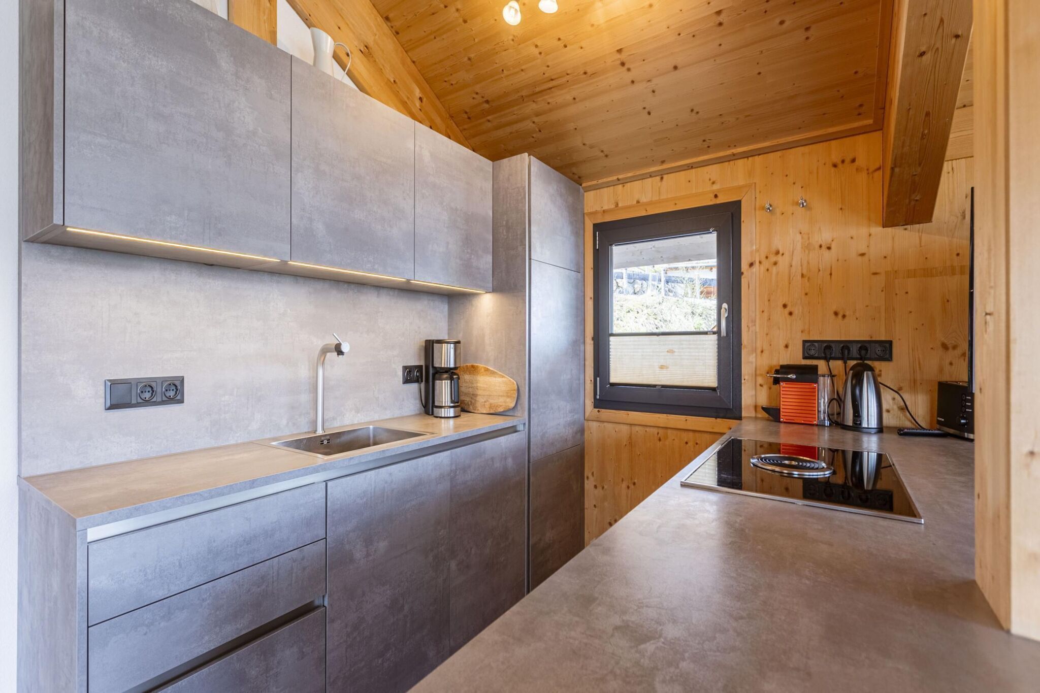 A modern kitchen with grey cabinets, a wooden ceiling, and a window providing natural light.