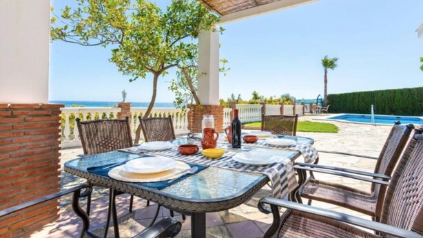 Een terras buiten met een eettafel, omringd door wickerstoelen, een zwembad en uitzicht op zee.
