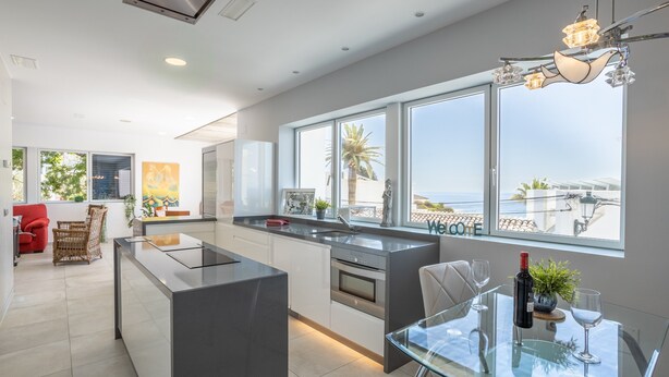 Une cuisine moderne dotée d’un grand îlot central, d’un coin repas avec une table en verre et d’une vue sur la mer.
