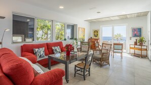 Un salon moderne avec un canapé rouge, une table à manger en verre, des chaises en osier et une vue sur la mer grâce à de grandes fenêtres.