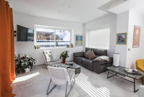 A modern living room with a sofa, armchair, and coffee table, a flat-screen TV, and a window with blinds.