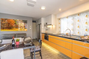 Cozinha moderna com armários laranja, área de jantar com sofá e uma grande pintura com tema de outono na parede.