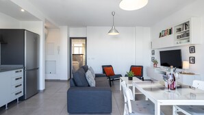 Una cocina moderna con refrigerador empotrado, un área de comedor con una mesa y sillas blancas, un sofá gris con cojines y un televisor montado en la pared.