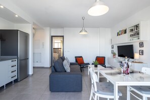 Une cuisine moderne dotée d’un réfrigérateur encastré, un coin repas avec une table et des chaises blanches, un canapé gris avec des coussins et un téléviseur fixé au mur.
