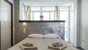 Un dormitorio con cama, almohadas, una mesita de noche, una ventana con cortinas y una repisa en la pared.